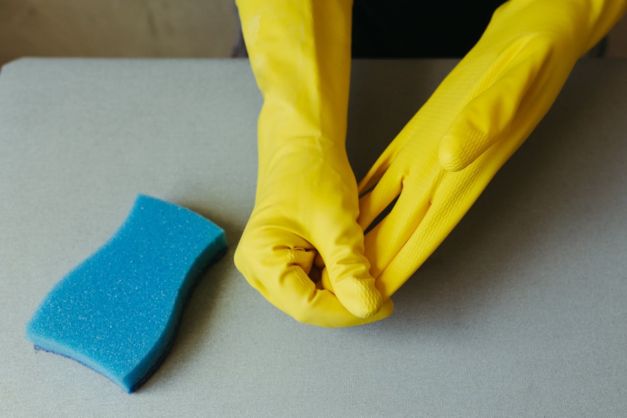 cta-bg Close-up of hands wearing yellow gloves next to blue sponge on gray surface.