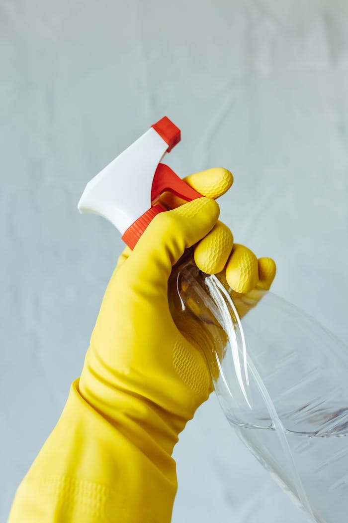 about-01 A hand wearing a yellow rubber glove holds a red and white spray bottle against a neutral background.