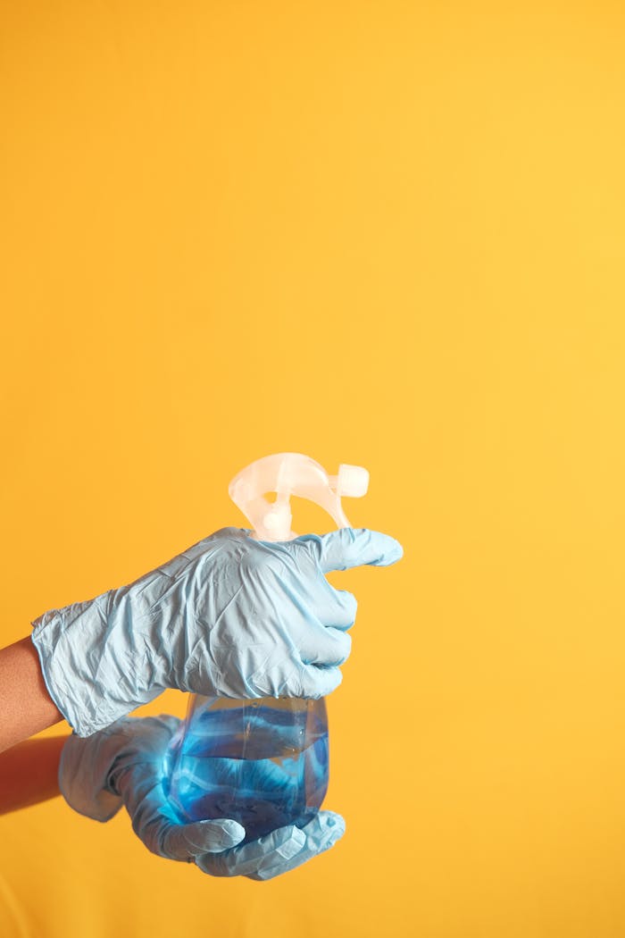 home-services-01 Hand in blue glove holding a cleaning spray bottle against a yellow background.
